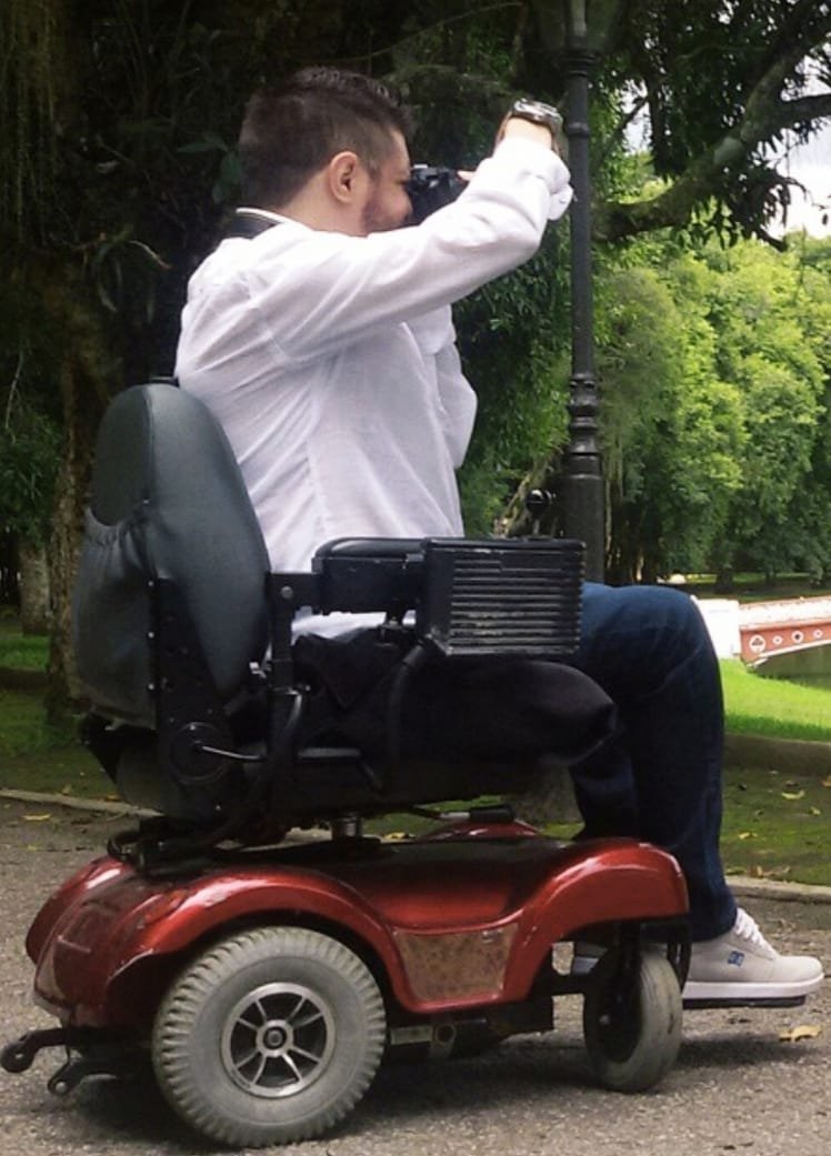 Berguim fotografando ao ar livre, em sua cadeira de rodas motorizada, junto a um poste em um parque.